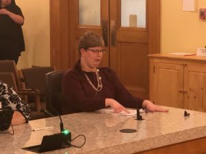 Chrissy Krumm sits in a hearing room, giving her testimony. She sits at a long table and has a microphone in front of her.